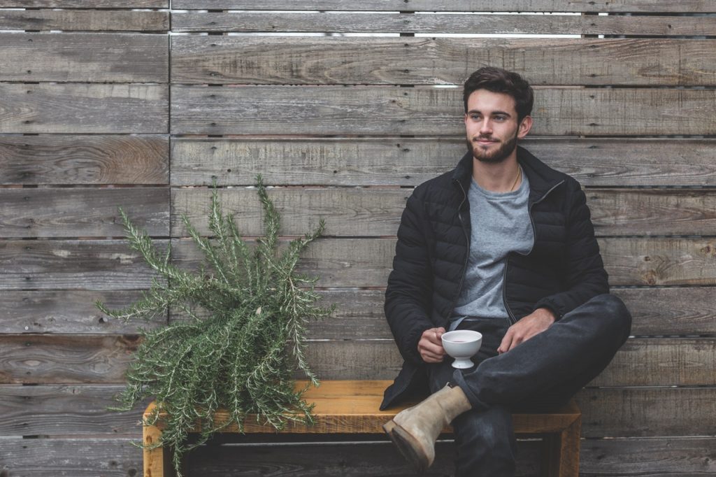 jeune homme assis sur un banc à côté d'une plante portant un t-shirt gris un jean des bottines beiges et une doudoune noire