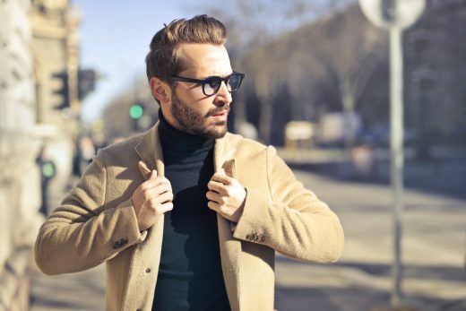 homme portant des lunettes, un pull à col roulé noir et une veste beige